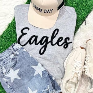 May include: A light gray t-shirt with the black text Eagles, a beige and gray baseball cap with the text GAME DAY, light blue denim shorts with a white star pattern, and a pair of off-white athletic sneakers with white laces and soles.