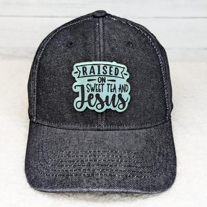 May include: A dark denim baseball cap with a curved brim. The cap features a rectangular patch with the words "RAISED ON SWEET TEA AND Jesus" in a teal color. The hat is a classic six-panel design.