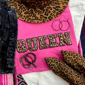May include: A vibrant pink t-shirt with the word "QUEEN" in leopard print lettering. The image also features a leopard print hat, hoop earrings, bracelets, and leopard print shoes. A white denim jacket and a black jacket are also visible.
