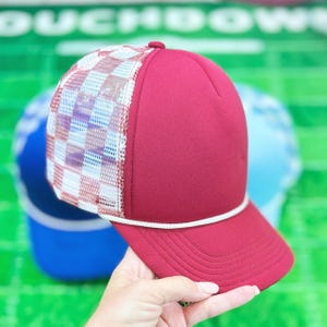 May include: A burgundy trucker hat with a white and checkered mesh panel. The hat has a white rope detail and a curved brim. The background features other hats and a green surface.