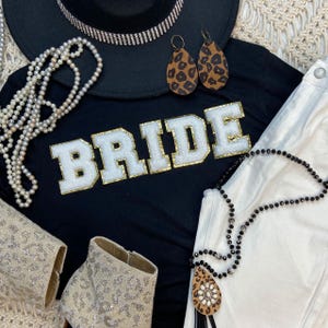 May include: A black t-shirt with the word "BRIDE" in white and gold lettering, paired with white pants, a black hat with rhinestones, leopard print earrings, and beaded necklaces. Also includes a pair of sparkly boots.