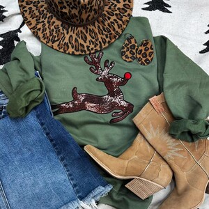 May include: An olive green sweatshirt with a sequined reindeer design, paired with denim shorts, tan cowboy boots, and a leopard print hat and earrings. The reindeer has a red nose. The outfit is arranged on a patterned white and black surface.