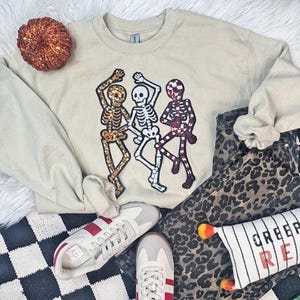 May include: Beige sweatshirt with three dancing skeleton figures. The skeletons are embellished with gold, white, and burgundy sequins. The image also includes a pair of sneakers, leopard print pants, and a decorative pumpkin.