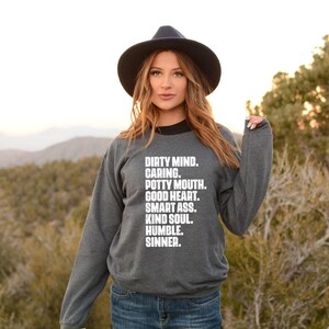 May include: A dark gray sweatshirt with the text "DIRTY MIND. CARING. POTTY MOUTH. GOOD HEART. SMART ASS. KIND SOUL. HUMBLE. SINNER." in white. The sweatshirt has a black collar and is worn with blue jeans and a black hat.