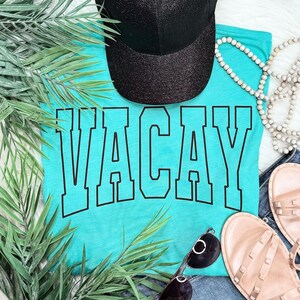 May include: A turquoise t-shirt with the word "VACAY" in large black block letters. A black glitter baseball cap sits above the shirt. Sunglasses, sandals, and a beaded necklace are also visible.