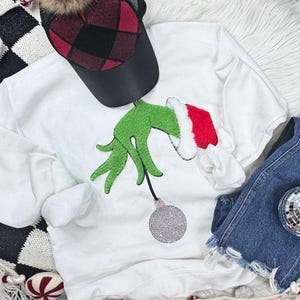 May include: White sweatshirt featuring a green hand with a red and white Santa hat, holding a rhinestone-covered ornament. A black and red plaid hat and denim shorts are also in the image. The sweatshirt is a festive holiday design.