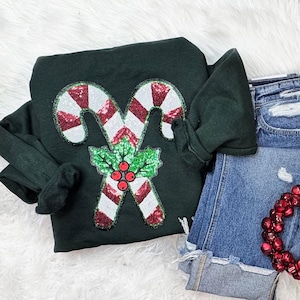 May include: Dark green sweatshirt featuring a sequined candy cane design with red and white stripes, green holly leaves, and red berries. The sweatshirt is paired with denim shorts and a red beaded garland, creating a festive holiday look.