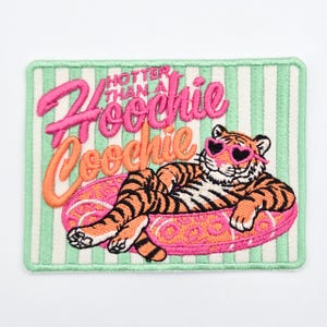 May include: A rectangular embroidered patch featuring a light green and white striped background. The text "HOTTER THAN A Hoochie Coochie" is stitched in pink and orange. A tiger wearing pink heart-shaped sunglasses lounges on a pink leopard print float.