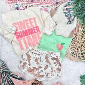 May include: A cream-colored sweatshirt with the text "SWEET SUMMER TIME" in pink, paired with a light green top with a peach design, cow-print shorts, and white ankle boots. The items are arranged on a white fluffy surface.