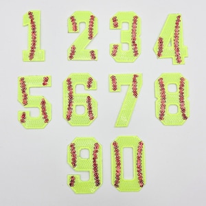 May include: A collection of neon yellow sequined number patches, from 1 to 0. Each number is outlined with red sequins, resembling baseball stitching. The numbers are arranged on a white background.