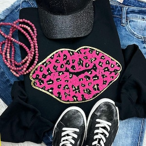 May include: A black sweatshirt with a pink leopard print lip design, a black glitter baseball cap, a pink beaded necklace, black sneakers, and blue jeans. The items are arranged on a white surface.