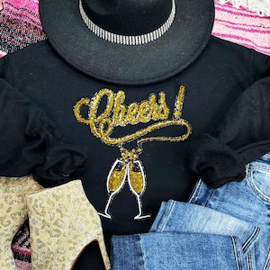 May include: Black sweatshirt with gold glitter "Cheers!" text and champagne glasses graphic. A black hat with a silver band, denim jeans, and gold patterned boots are also in the image.