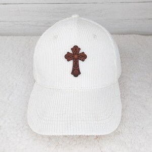 May include: A white corduroy baseball cap featuring a decorative brown cross emblem on the front. The cap has a curved brim and a structured crown. The cross design has a detailed, patterned appearance.