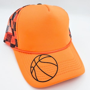 May include: Bright orange trucker hat with a black basketball graphic on the front. The hat features a mesh back with a black and red checkered pattern. A black rope detail runs along the brim.