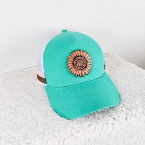 May include: A turquoise baseball cap with a wooden sunflower applique. The cap features a white mesh back, a curved brim, and a brown and pink accent stripe. The sunflower is a detailed, laser-cut design.
