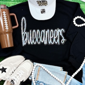 May include: A black long-sleeve shirt with the word "Buccaneers" in sequined lettering. The image also includes a white visor with "Game Day" text, a brown football-shaped tumbler, white sneakers, and a beaded bracelet.