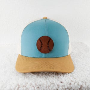 May include: A baseball cap with a light blue front panel, tan brim, and white mesh back. A brown leather baseball patch is centered on the front. The cap has a curved brim and a button on top.