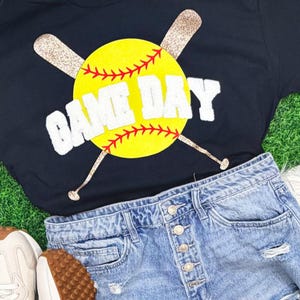 May include: A navy blue t-shirt with a yellow softball design, red stitching, and the words "GAME DAY" in white. Two glittery baseball bats cross behind the softball. Light blue denim shorts and white sneakers complete the sporty look.