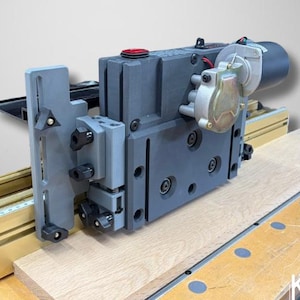 May include: A gray and black woodworking machine with a red button, mounted on a wooden surface. The machine has a motor and is designed for precision cutting. The brand name "KEKAP DESIGNS" is visible.