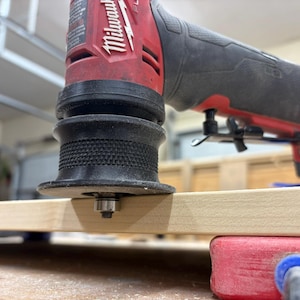 May include: A red and black Milwaukee oscillating multi-tool is positioned over a light-colored wooden board. The tool's black base is in contact with the wood, and a red clamp secures the board. The tool is designed for woodworking and DIY projects.