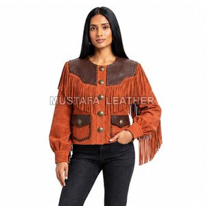 Puede incluir: Una chaqueta de ante color óxido con canesú de cuero marrón oscuro, largos flecos en el pecho y las mangas, y botones metálicos. La chaqueta incluye dos bolsillos delanteros con solapa y el texto MUSTAFA LEATHER.