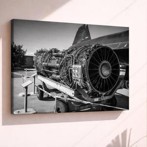 May include: Black and white canvas print of a jet engine on a wheeled cart. The engine is large, with intricate details of pipes and machinery. The background includes a plane's tail and a sunny outdoor setting.