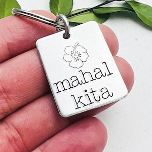 Puede incluir: Llavero de plata con una flor de hibisco y el texto "mahal kita", que significa "te quiero" en tagalo.