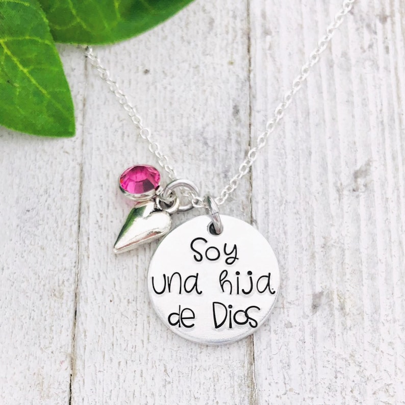 May include: Silver necklace with a round pendant that says "Soy una hija de Dios" in Spanish. The pendant is attached to a small silver heart and a pink crystal.