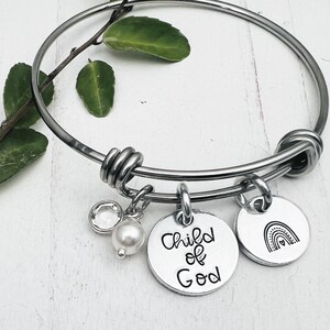 Pode incluir: Pulseira de bracelete de prata com pingentes. Os pingentes incluem uma pérola, um cristal transparente, um pingente com o texto "Filho de Deus" e um pingente com um design de arco-íris.