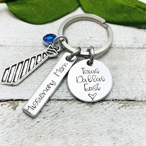May include: A silver keychain with a tie charm, a rectangular charm with "Missionary Mom" engraved, and a round charm with "Texas Dallas East" and a heart. A blue gem is attached.