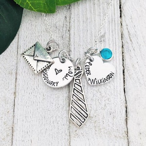 Puede incluir: Collar de plata con varios colgantes, incluyendo un corazón con la inscripción "Mamá", una corbata, un sobre y un corazón con la inscripción "Elder Wilkinson". También se incluye una piedra preciosa azul turquesa.