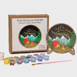 May include: A paint-by-layers craft kit with a shadow box design. The kit includes a cardboard box with the text "Paint By Layers Craft Kit Summer Nights Shadow Box". The shadow box features a camping scene with a red tent, campfire, and mountains. Paint pots and a paintbrush are included.