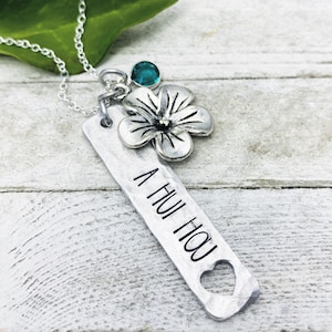 May include: Silver necklace with a silver flower charm and a teal blue gemstone. The pendant has the words "A Hui Hou" engraved on it.
