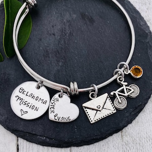 Puede incluir: Una pulsera de brazalete plateada con múltiples dijes. Los dijes incluyen un colgante redondo con el texto "Oklahoma Mission", un corazón con "Sister Evans", un sobre, una bicicleta y una pequeña gema naranja. La pulsera está sobre una superficie oscura.