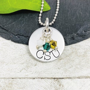 May include: A silver-colored pendant necklace with a ball chain. The round pendant has the letters "CSU" engraved on it. Three small, colorful crystals in white, green, and gold hang from the pendant.