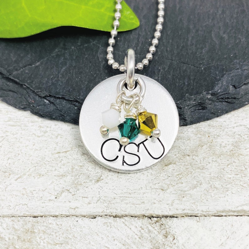 May include: A silver-colored pendant necklace with a ball chain. The round pendant has the letters "CSU" engraved on it. Three small, colorful crystals in white, green, and gold hang from the pendant.