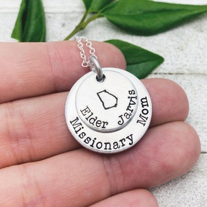 Puede incluir: Collar de plata con un colgante de doble cara. La parte superior presenta el contorno del estado de Georgia y el texto "Jarvis Mom". La parte inferior presenta el texto "Elder Missionary".