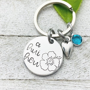 May include: A silver keychain with a round charm that says "a hui hou" with a hibiscus flower design. The keychain also has a silver heart charm and a blue crystal charm.