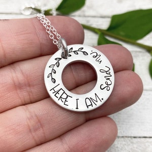 May include: A silver-colored pendant necklace with a circular design. The pendant has a central hole and is inscribed with the words "HERE I AM. Send me." A decorative leaf design is on the top left. The necklace is on a silver chain.