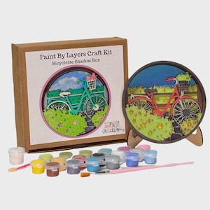 May include: A craft kit for a bicycle shadow box. The kit includes a cardboard box with the text "Paint By Layers Craft Kit Bicyclette Shadow Box", a painted bicycle shadow box, paint pots, and a paintbrush. The shadow box features a bicycle with a basket of flowers.