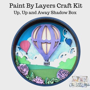 May include: A layered craft kit titled "Up, Up and Away Shadow Box." The circular design features a purple hot air balloon, pink and blue balloons, and floral accents. The background includes a blue sky and green landscape. The text "Paint By Layers Craft Kit" is at the top.