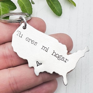 May include: A silver-colored keychain in the shape of the United States. The keychain has the Spanish text "Tu eres mi hogar" and a small heart symbol. The keychain is attached to a silver ring.