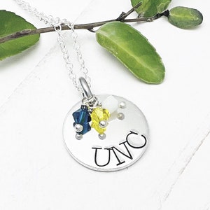 May include: A silver necklace with a round pendant. The pendant has the letters "UNC" stamped on it, with blue and yellow crystal beads. The necklace is on a white surface with green leaves.