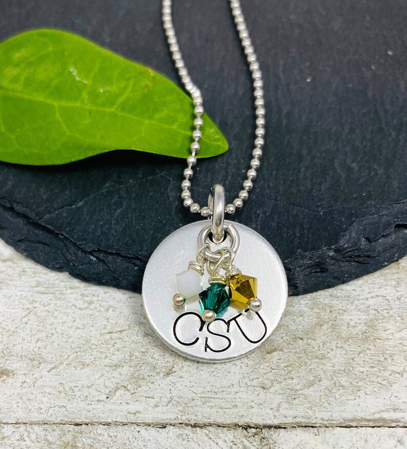 May include: A silver pendant necklace with a round charm. The charm has the letters "CSU" stamped on it, with three small, colorful crystals hanging below. The necklace chain is a ball chain.