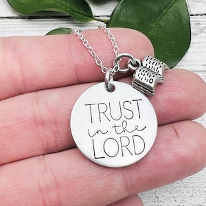 Peut inclure: Un collier argenté avec un pendentif rond portant l'inscription "TRUST in the LORD" en noir. Le collier est orné d'un charm en forme de deux petits livres. La chaîne est fine et le pendentif est circulaire.