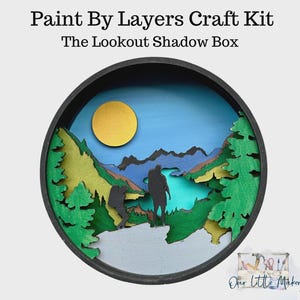 May include: A round, layered craft kit titled "Paint By Layers Craft Kit" and "The Lookout Shadow Box." The kit features a scenic landscape with a yellow sun, mountains, and two silhouetted figures. The frame is black.