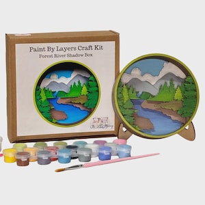 May include: A paint-by-layers craft kit with a forest river shadow box design. The kit includes a cardboard box, a wooden frame, paint pots, and a pink paintbrush. The shadow box features a layered landscape with mountains, trees, and a river.