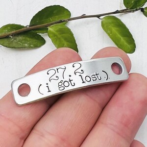 May include: A small, rectangular, silver-colored metal tag with rounded ends and two holes. The tag has the text "27.2 (i got lost)." stamped in a black, handwritten-style font. The tag is held in a person's hand.