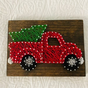 May include: Red truck with a green Christmas tree in the bed, made with string art on a wooden background.