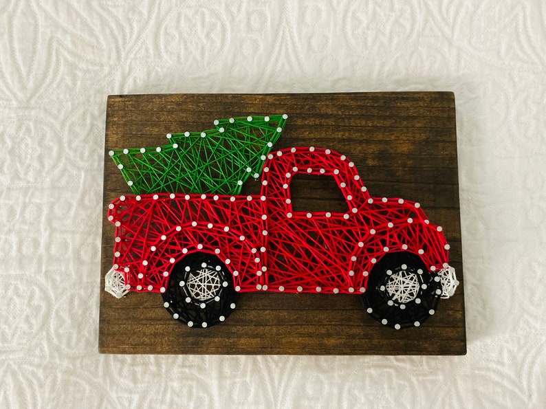 May include: Red truck with a green Christmas tree in the bed, made with string art on a wooden background.